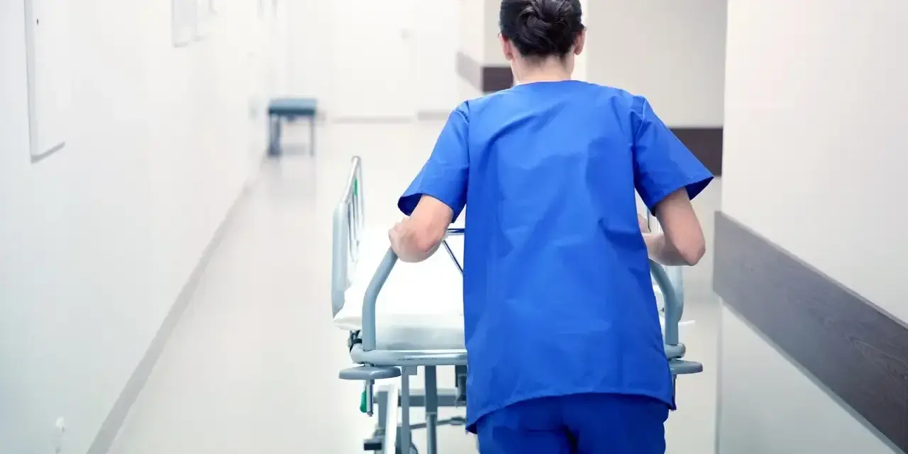 Salowa w niebieskim uniformie pcha łóżko szpitalne korytarzem. To część jej obowiązków w szpitalu.