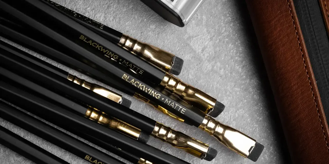 En samling svarta Blackwing-pennor med guldfattningar och suddgummin. En brun läderfodral och en metallbehållare syns också.