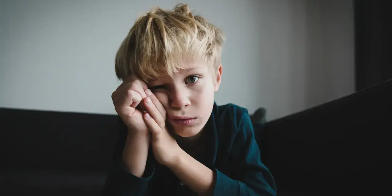Chłopiec z blond włosami, zamyślony, opiera głowę na dłoniach. Może to być obraz przedstawiający zespół Aspergera – objawy u nastolatków, takie jak trudności w komunikacji.