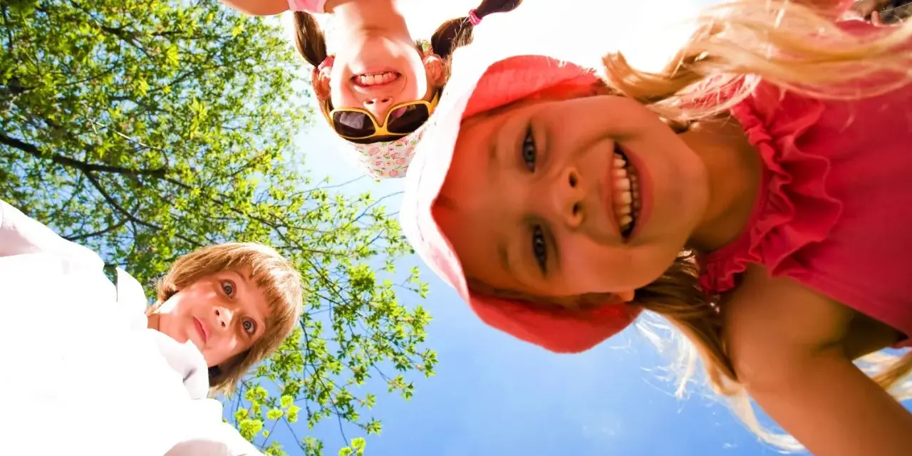 Trois enfants souriants, vus d'en bas, profitent d'une journée ensoleillée lors d'un camp sportif.