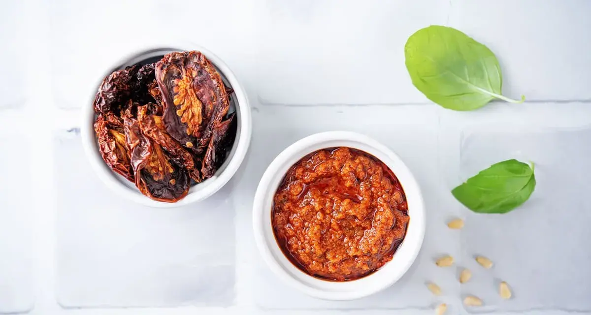 Due ciotoline bianche: una con pomodori secchi interi, l'altra con un invitante pesto di pomodori secchi. Foglie di basilico e pinoli completano la scena.