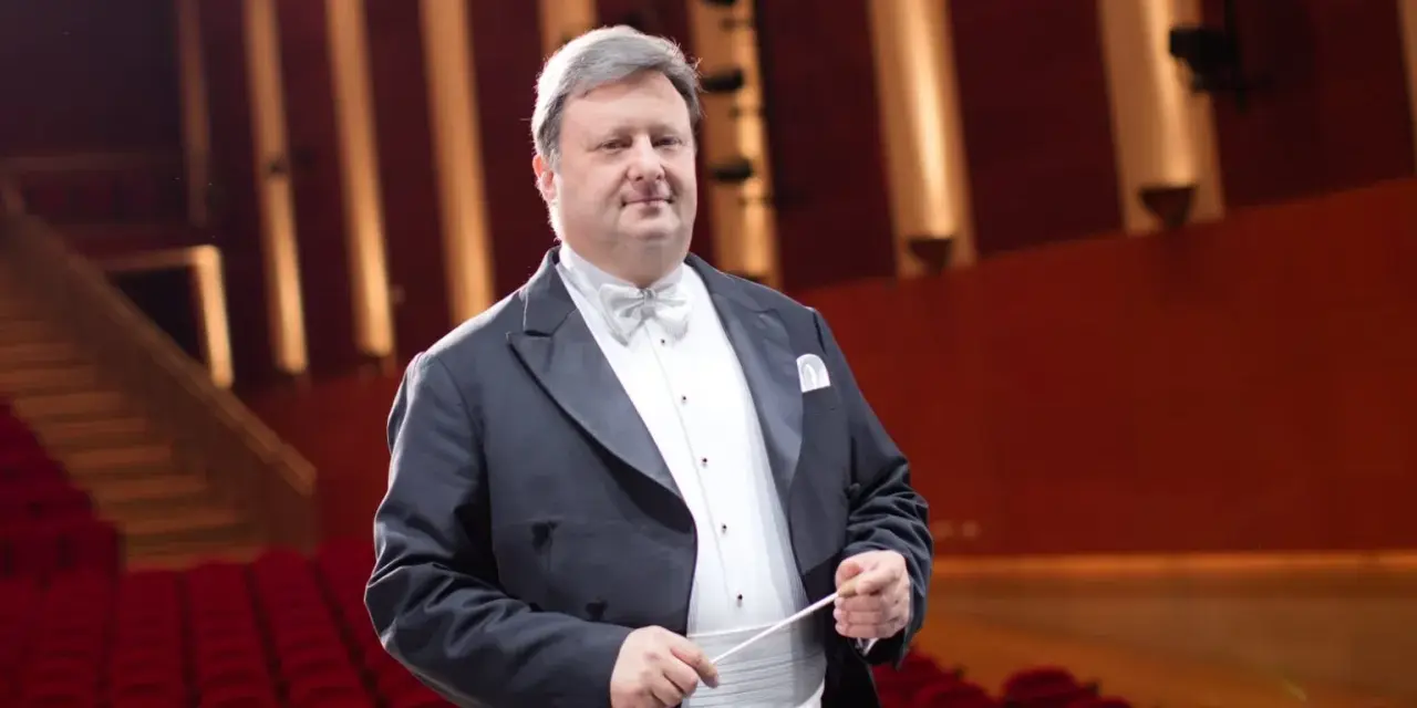 Jacek Boniecki, dyrygent w eleganckim fraku, z batutą w dłoni, na tle czerwonej widowni. Jego życiorys to historia pasji do muzyki.