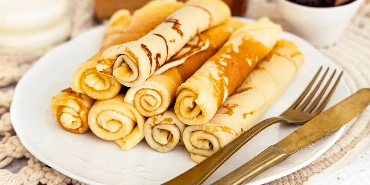 Ein Teller mit goldbraunen Quark Pfannkuchen, aufgerollt und gestapelt. Daneben liegen ein goldenes Messer und eine Gabel. Im Hintergrund eine Schale Marmelade.