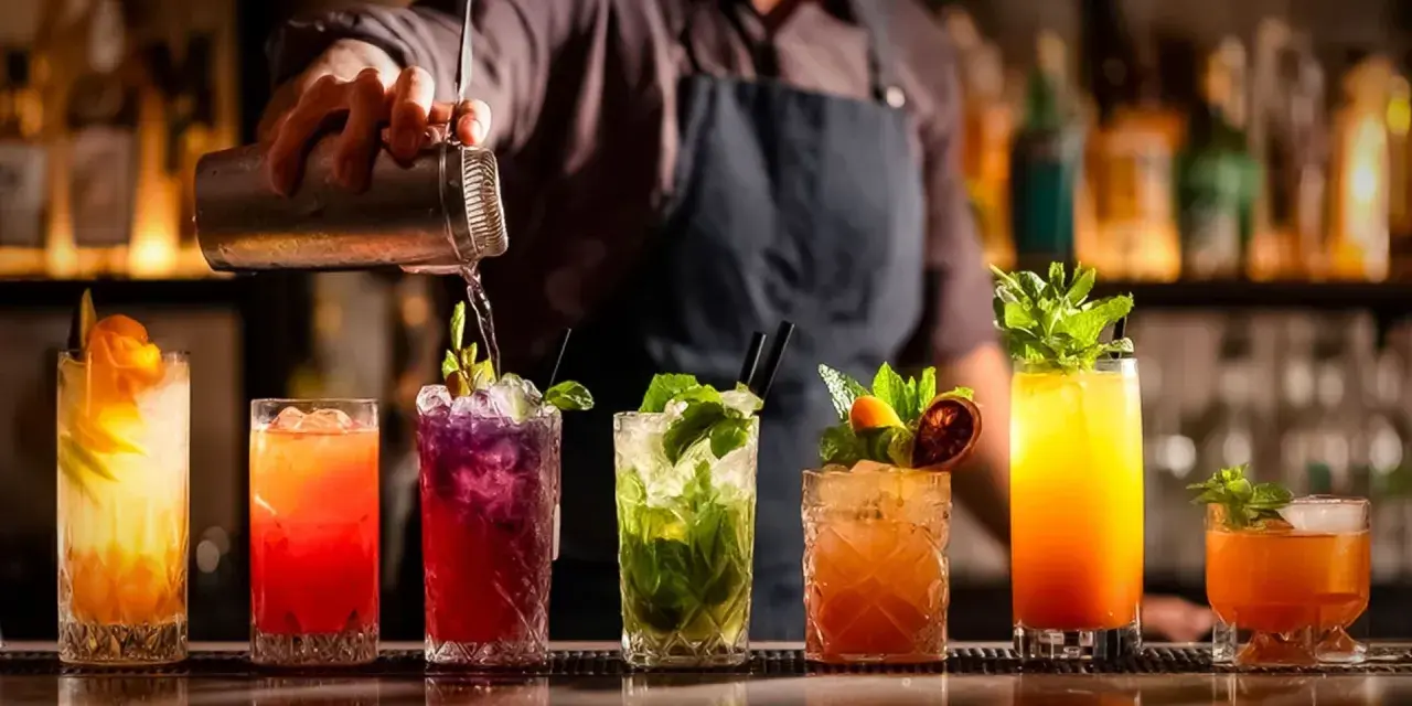 Barman przelewa drinka z shakera. Na blacie stoi rząd kolorowych koktajli, każdy ozdobiony owocami i miętą. Idealny przykład, jak zrobić dobry drink.