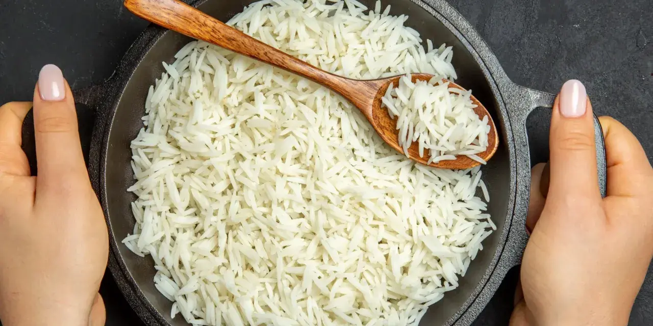 Dwie ręce trzymają okrągłą, czarną patelnię pełną puszystego ryżu basmati. Drewniana łyżka zanurzona w ryżu.