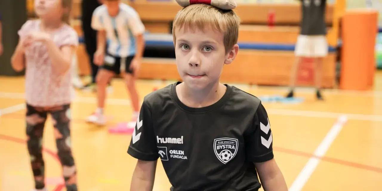 Chłopiec z zabawką na głowie, w koszulce KS Bysstra, podczas zajęć sportowych.
