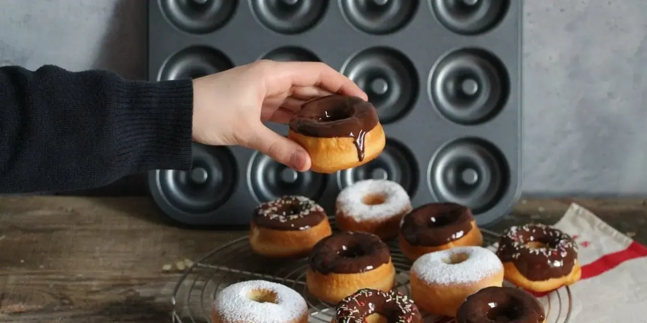 Une main tient un donut glacé au chocolat, prêt à être dégusté. Une belle collection de donuts au four, certains saupoudrés de sucre glace, d'autres de glaçage au chocolat et de vermicelles colorés, attendent sur une grille.
