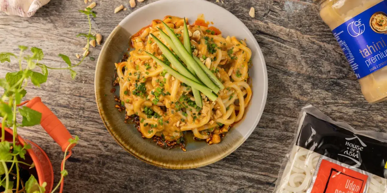 Makaron udon w sosie orzechowym, udekorowany ogórkiem i szczypiorkiem. Obok opakowanie makaronu udon mrożony czy suszony.