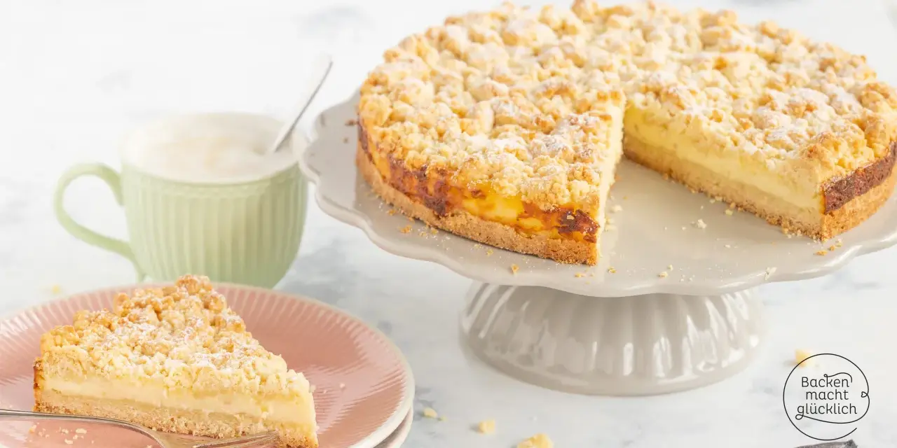 Ein saftiger Streuselkuchen mit Puddingfüllung, bestäubt mit Puderzucker, neben einer Tasse Cappuccino.