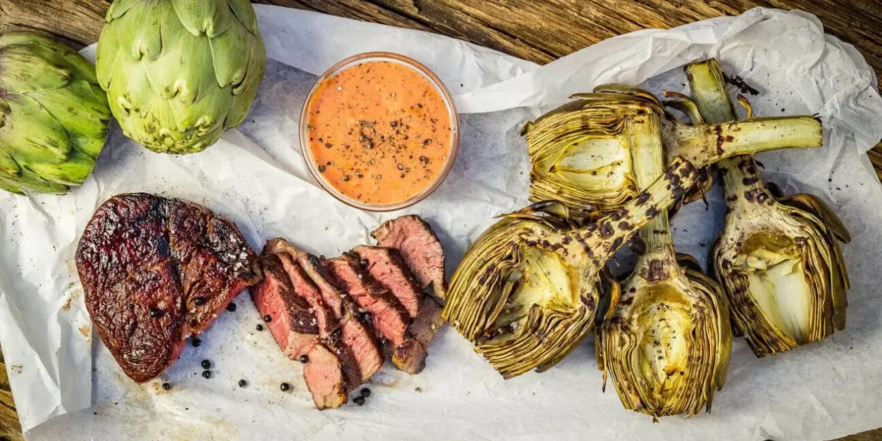 Saftiges Steak und gegrillte Artischocken mit Dip auf Holzuntergrund.