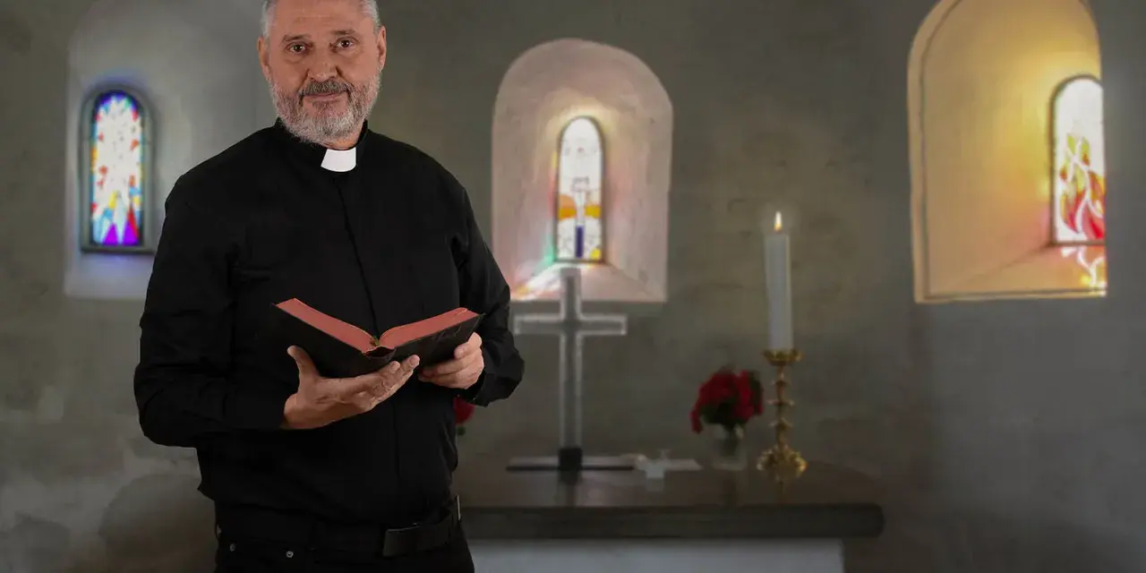 Ein Mann mit grauem Bart und Priesterkragen hält ein Buch in einer Kirche. Er hat sich entschieden, Priester zu werden.