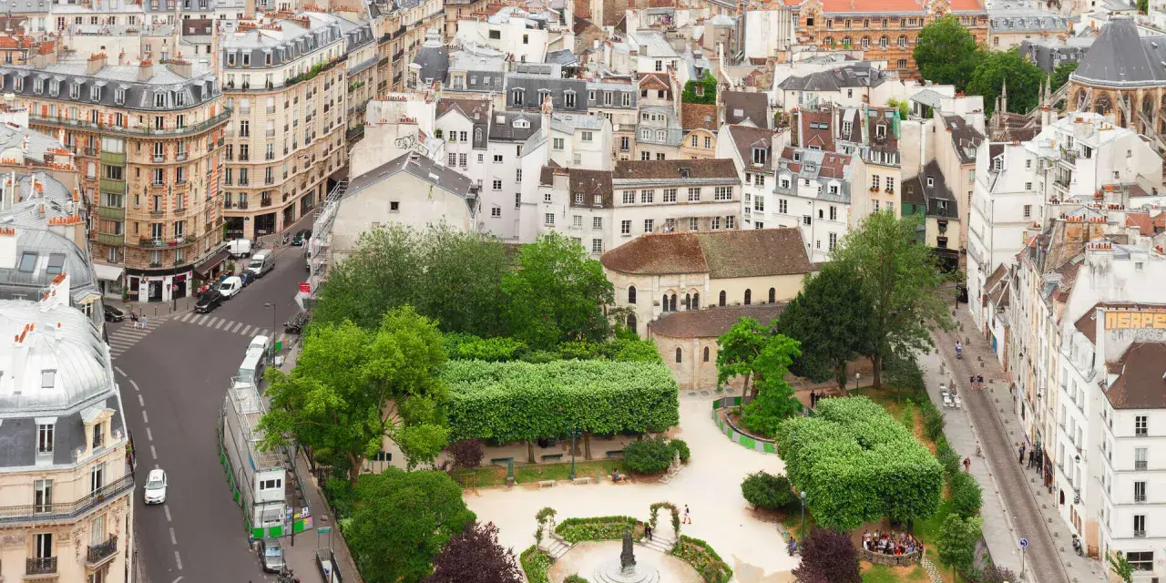 Vista aérea de un barrio latino con edificios antiguos, calles estrechas y un parque verde con gente disfrutando del día.