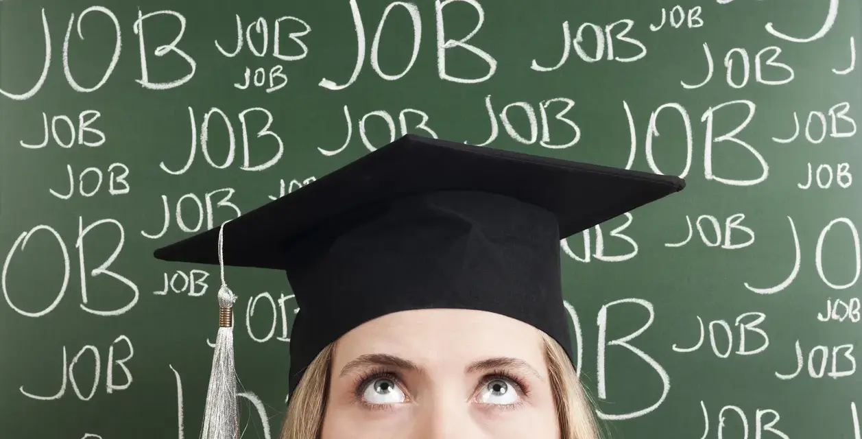 Absolventin mit Doktortitel blickt nach oben, während die Tafel voller "JOB" steht. Die Suche nach Arbeit nach dem Studium ist intensiv.