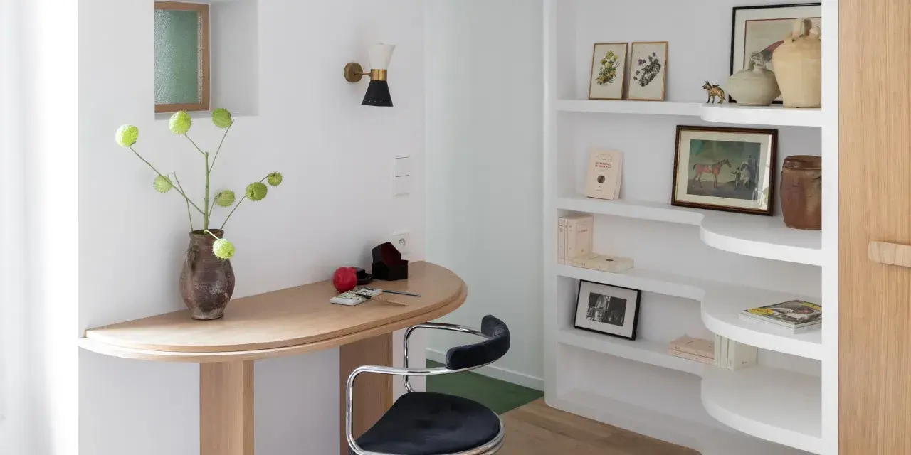 Un despacho pequeño en casa con un escritorio curvo de madera, una silla de terciopelo azul y estanterías blancas llenas de libros y objetos decorativos.