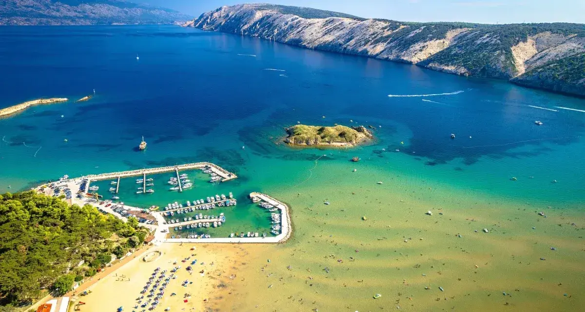 Piaszczyste plaże w Chorwacji, z turkusową wodą, łodziami w marinie i ludźmi cieszącymi się słońcem.