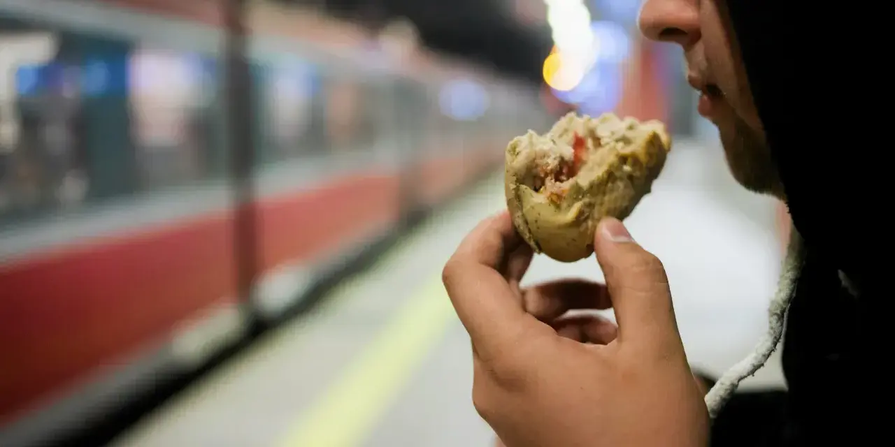 Osoba w kapturze je kanapkę na tle pociągu na peronie.