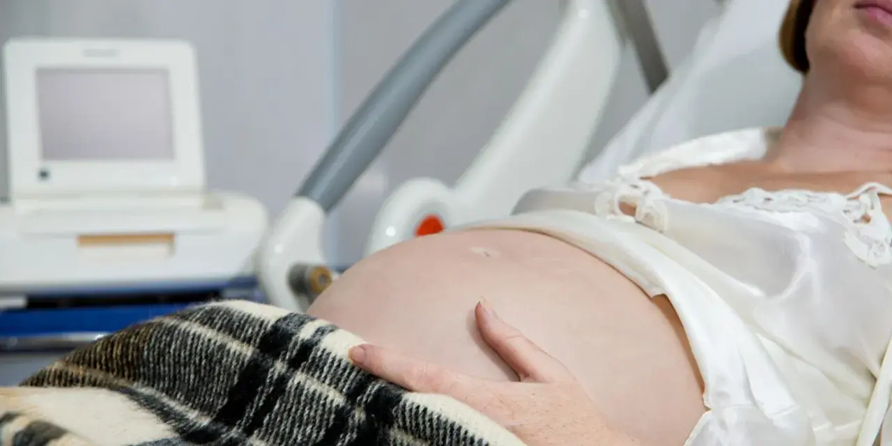 Brzuch kobiety w ciąży, ręka na brzuchu, monitor KTG i łóżko szpitalne.