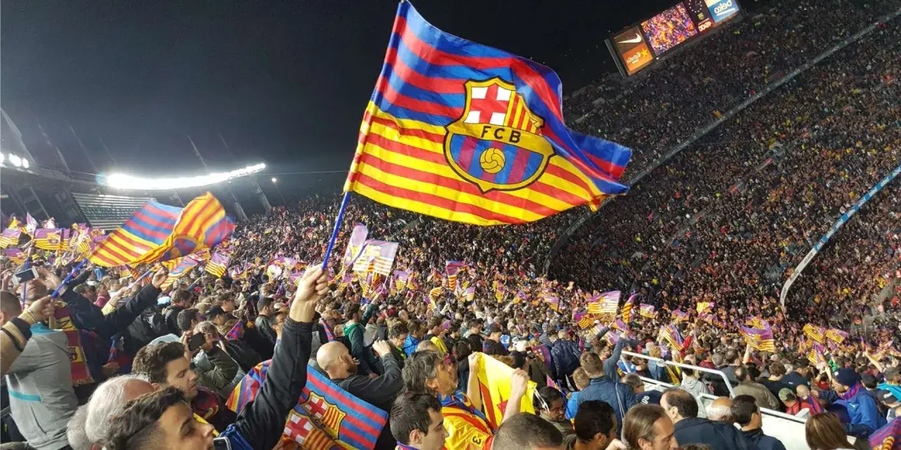 Tłum kibiców na stadionie unosi flagi z herbem FC Barcelona. Flaga Katalonii powiewa dumnie nad głowami fanów.