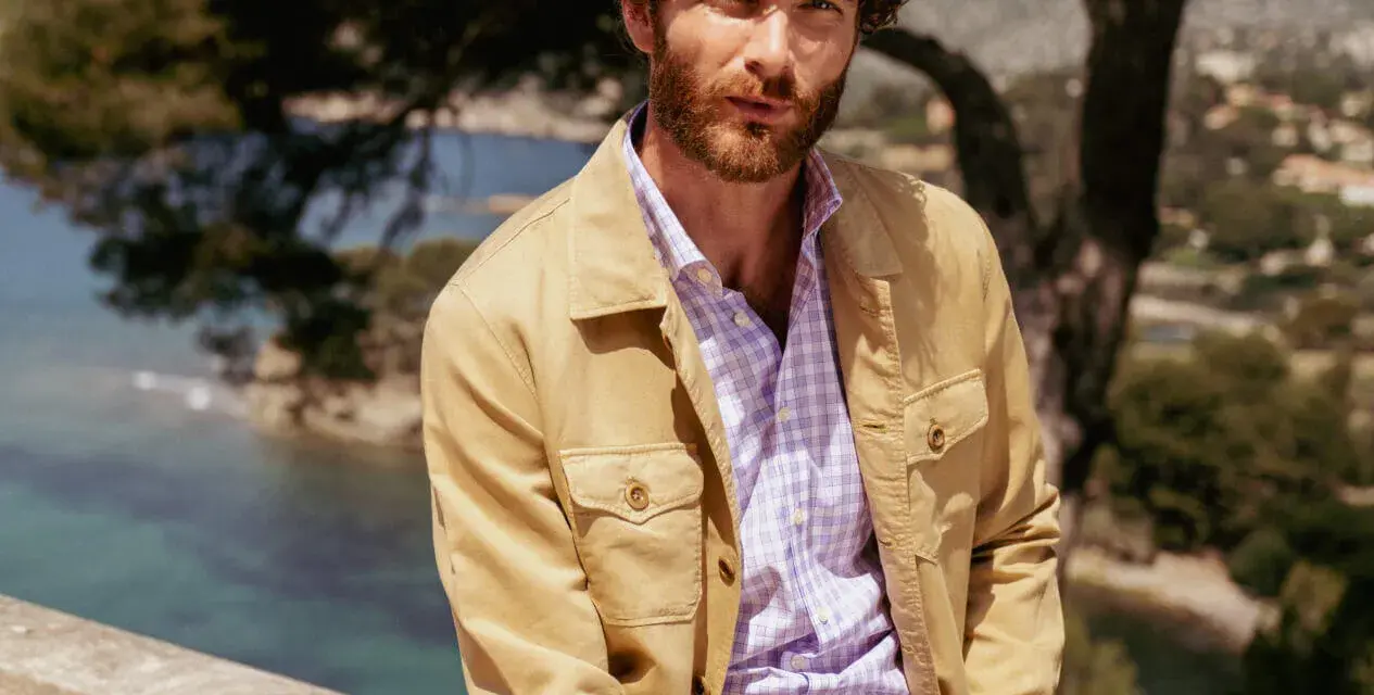 Un uomo con barba e capelli ricci indossa una camicia a quadri viola e una giacca color sabbia. Ottima idea su cosa mettere sopra la camicia uomo per un look casual chic.