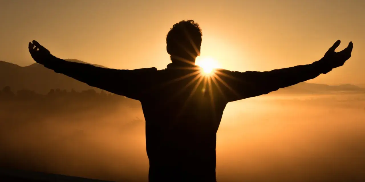 Silhouette eines Mannes mit ausgebreiteten Armen im Sonnenaufgang. Die Sonne bricht durch seine Silhouette und wirft Strahlen. Ein Symbol für Hoffnung und wie wird man Christ.