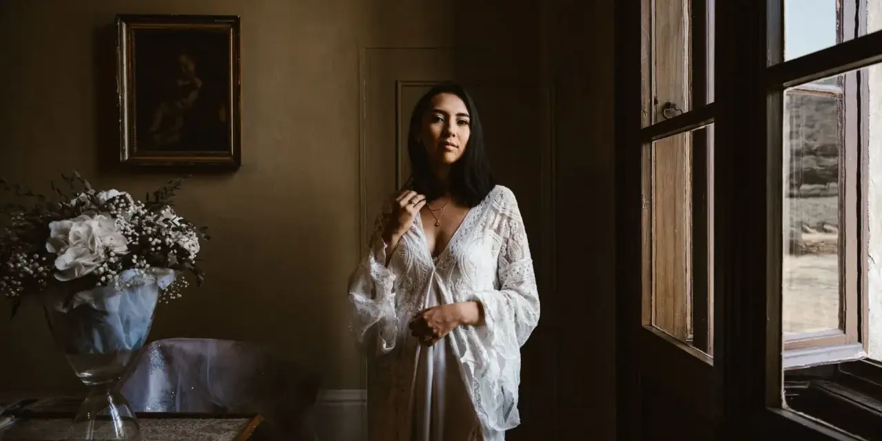 Kobieta w białej sukni, obok bukiet kwiatów. Scena sugeruje przygotowania do ślubu za granicą, co wyjaśnia przyczyny popularności wesel typu "destination wedding".