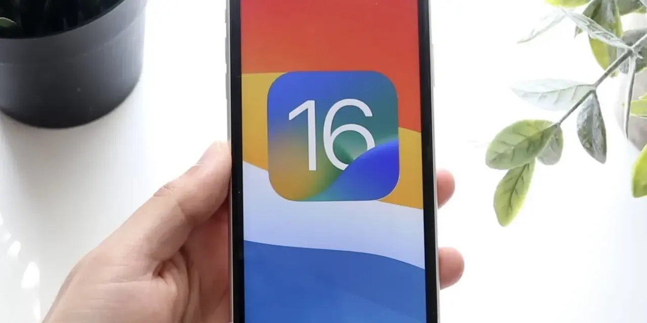 Dłoń trzyma iPhone'a z ikoną iOS 16 na kolorowej tapetcie.