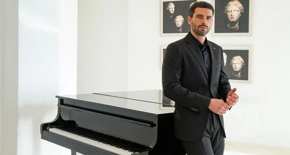Uomo con elegante outfit per andare a teatro, in completo nero, accanto a un pianoforte a coda.