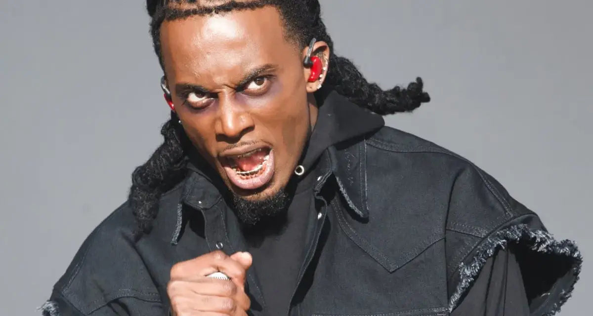 Playboi Carti, wiek nieznany, w czarnej kurtce z frędzlami, z mikrofonem w dłoni, z wyrazem twarzy pełnym pasji.