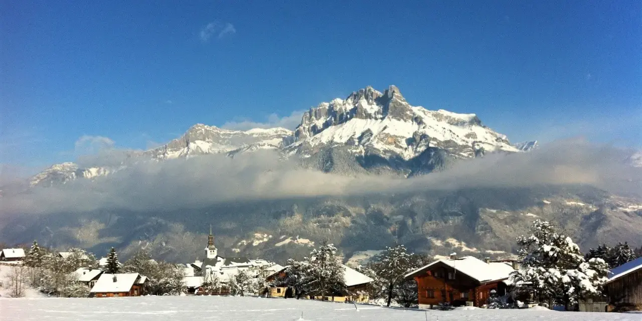 Un pueblo alpino cubierto de nieve, con casas de madera y una iglesia, bajo imponentes montañas nevadas. Un paisaje de invierno en francés.