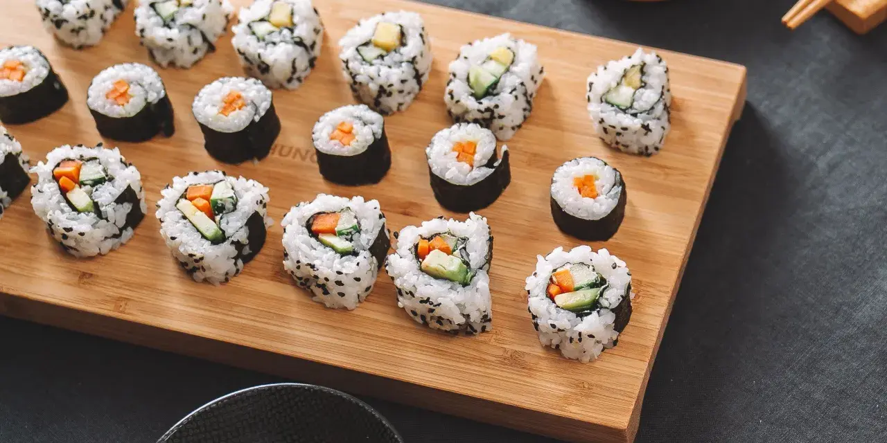 Frisch zubereitetes Sushi-Rezept auf einem Holzbrett, mit Gurke, Karotte und Ei gefüllt. Dazu Sojasauce und eingelegter Ingwer.