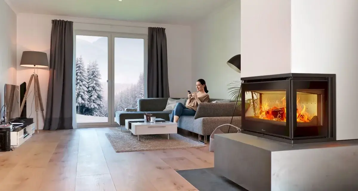 Mujer en sofá mira móvil junto a chimenea de cassette pellets con fuego vivo. Ventana muestra paisaje nevado.