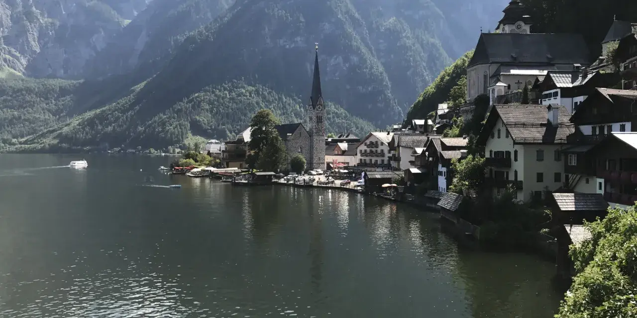 Malowniczy widok na Hallstatt, miasteczko nad jeziorem, z górami w tle. Widać kościół i domy, a na wodzie statek. To miejsce słynie z kopalni soli.