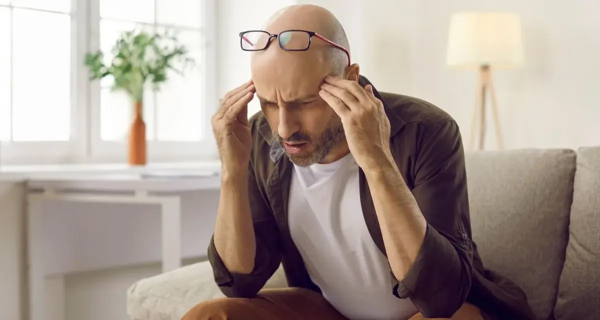 Mężczyzna z bólem głowy, być może objawem problemów z neuropsychologią, siedzi na kanapie, masując skronie.