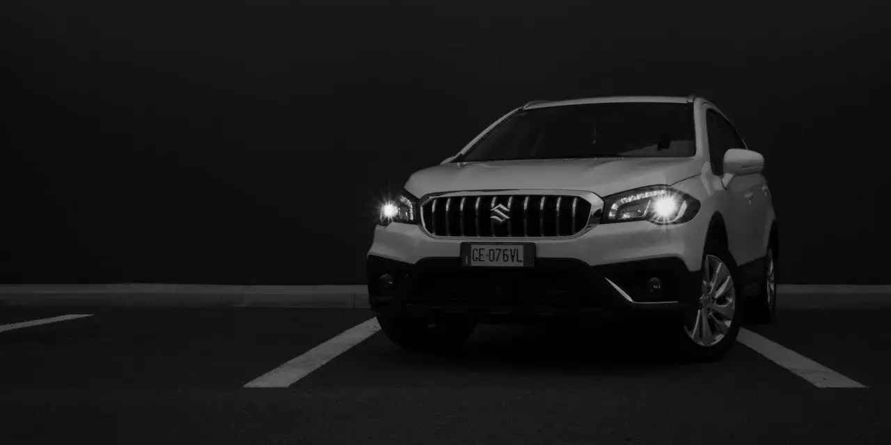 Białe Suzuki SX4 na ciemnym tle, z włączonymi światłami.