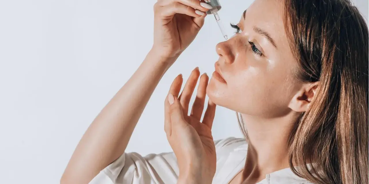 Frauenhand trägt Tropfen eines Serums auf ihr Gesicht auf. Ob Squalan oder Hyaluron, die Haut strahlt Feuchtigkeit aus.