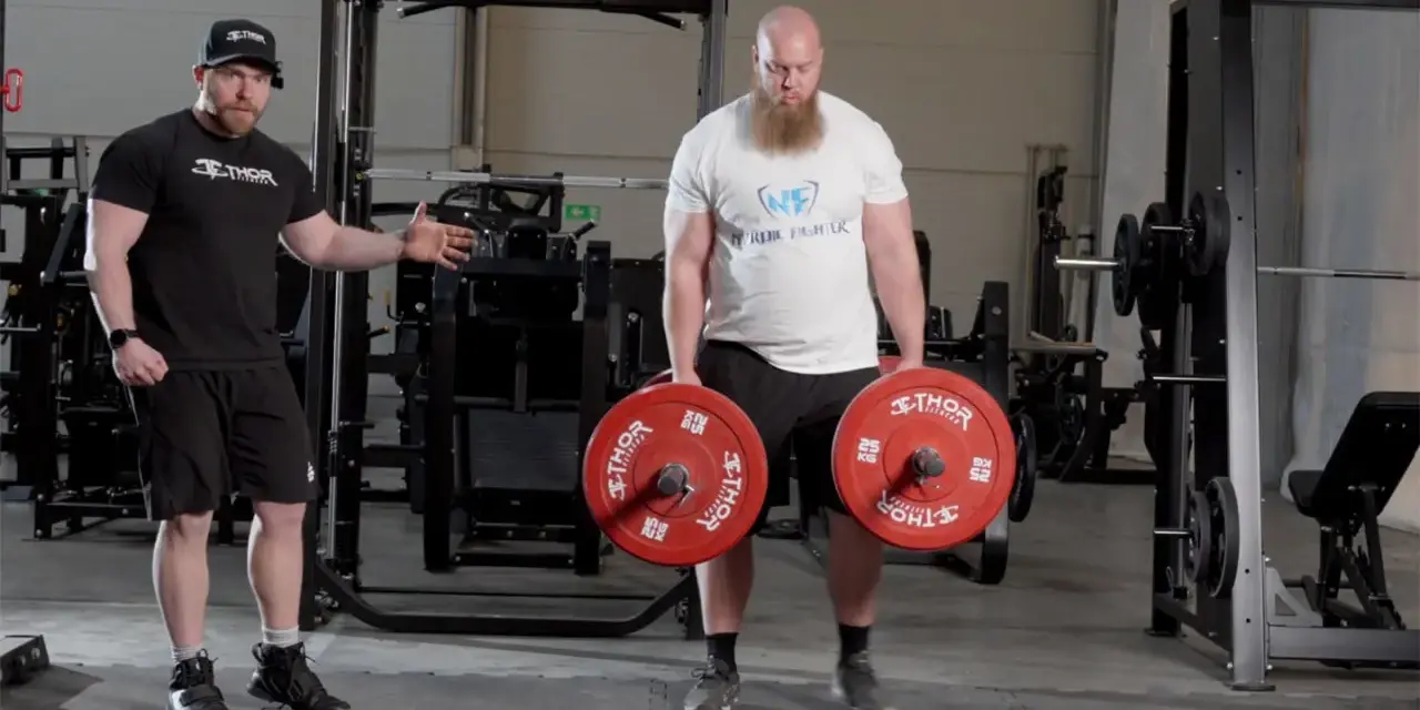 En man med skägg utför farmers walk med tunga vikter. En tränare visar hur övningen ska göras.