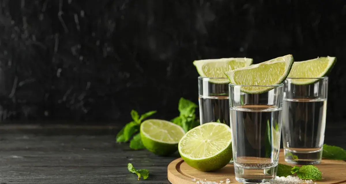 Trzy kieliszki z drinkiem z mezcalem lub tequilą, ozdobione ćwiartkami limonki i miętą, na tle czarnej ściany.