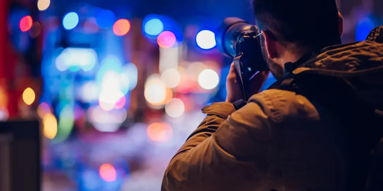 Mężczyzna z aparatem w ręku, uchwycony w trakcie nocnej fotografii. Rozmyte, kolorowe światła miasta tworzą magiczne tło.