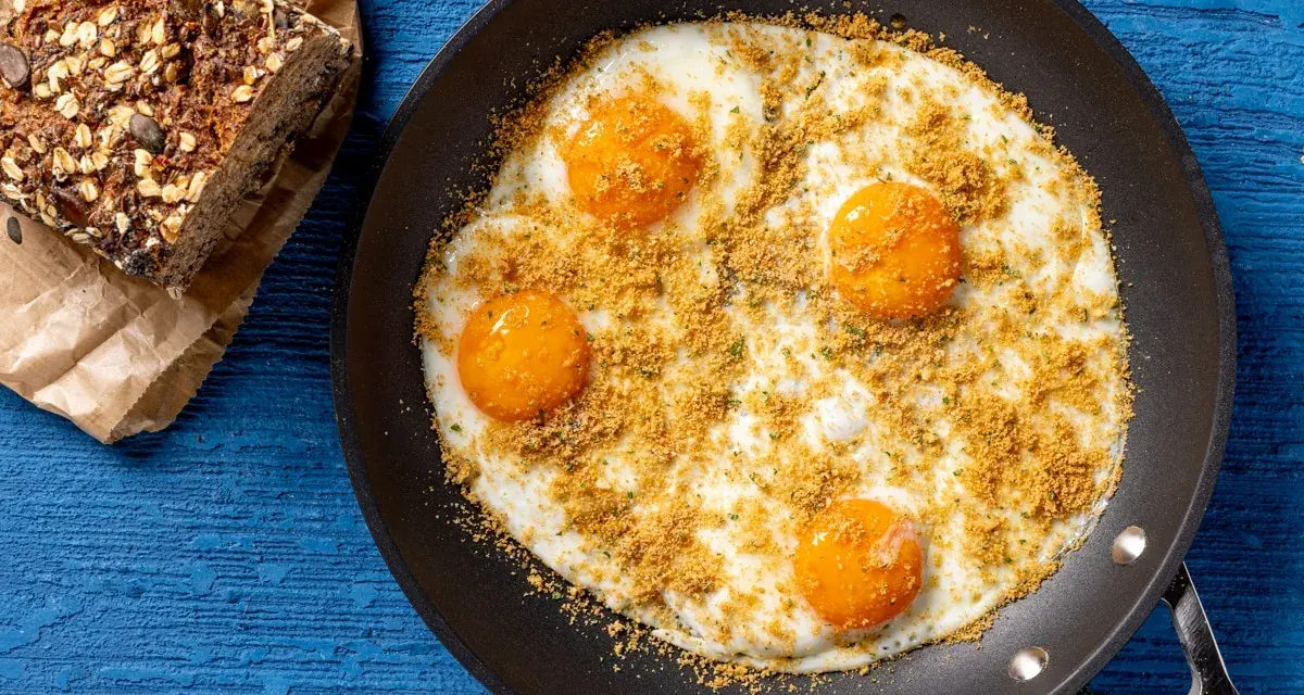 Uova fritte in padella, condite con pangrattato e erbe aromatiche, perfette per le tue ricette con uova. Accanto, una fetta di pane integrale.