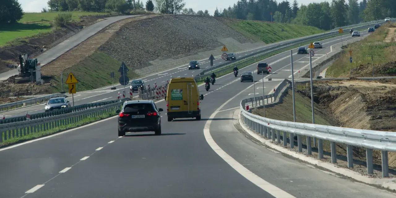 Droga ekspresowa z samochodami i motocyklami, gdzie panuje bezpieczna prędkość. W tle widać prace budowlane.