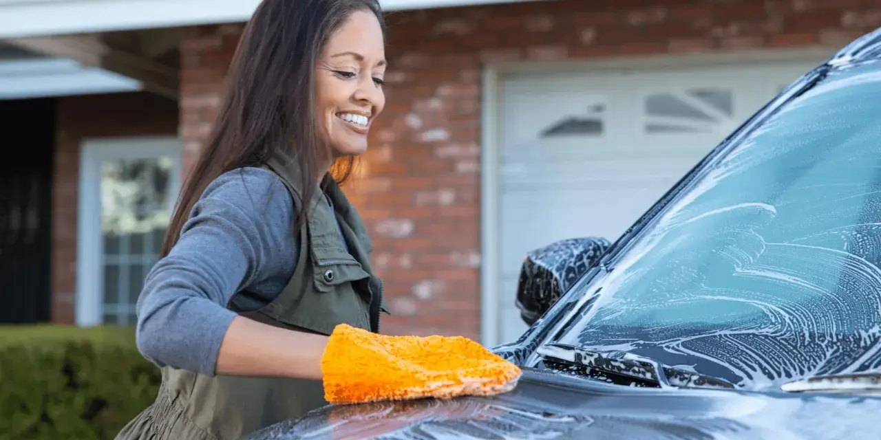 Glad kvinna tvättar bilen med en orange svamp. Hon putsar bilrutan som är täckt av skum.