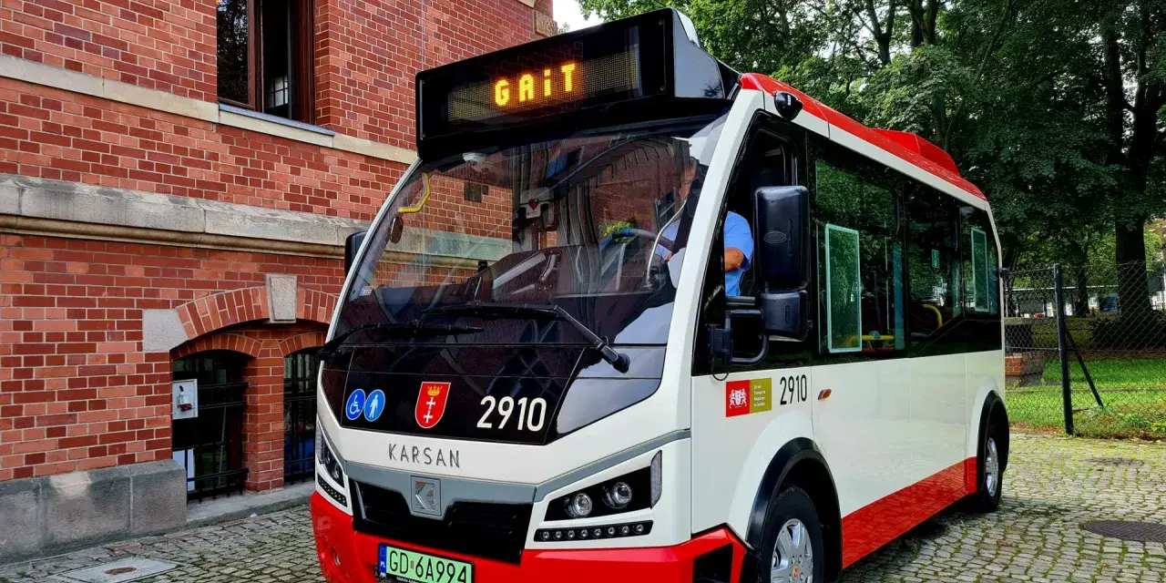 Nowy autobus 100 z napisem "GAIT" na wyświetlaczu, zaparkowany na brukowanej ulicy obok ceglanego budynku.