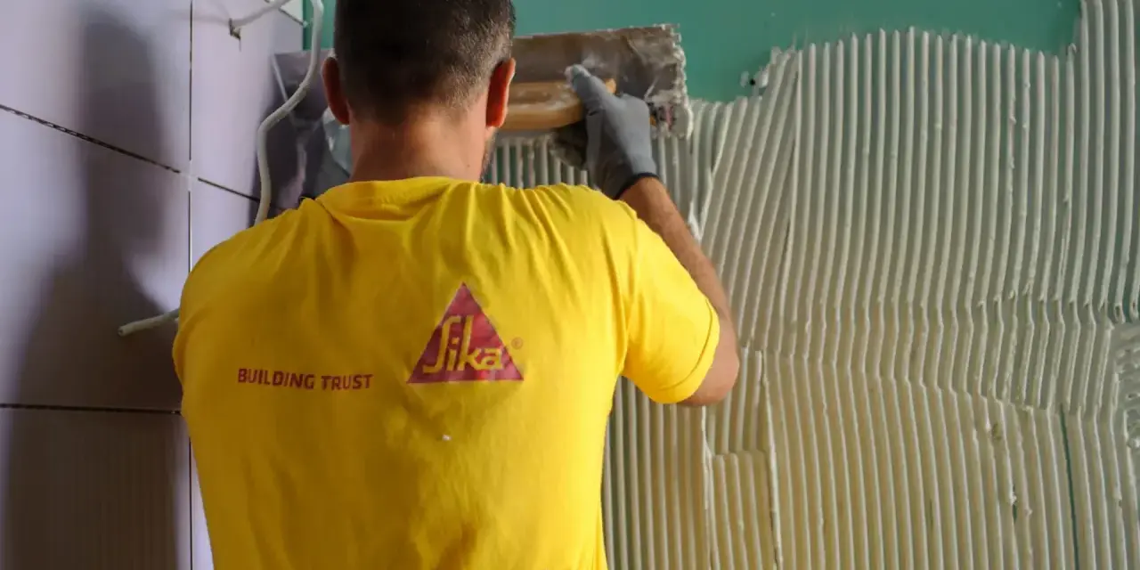 Un operaio con maglia gialla applica colla sul muro, mostrando come attaccare piastrelle.