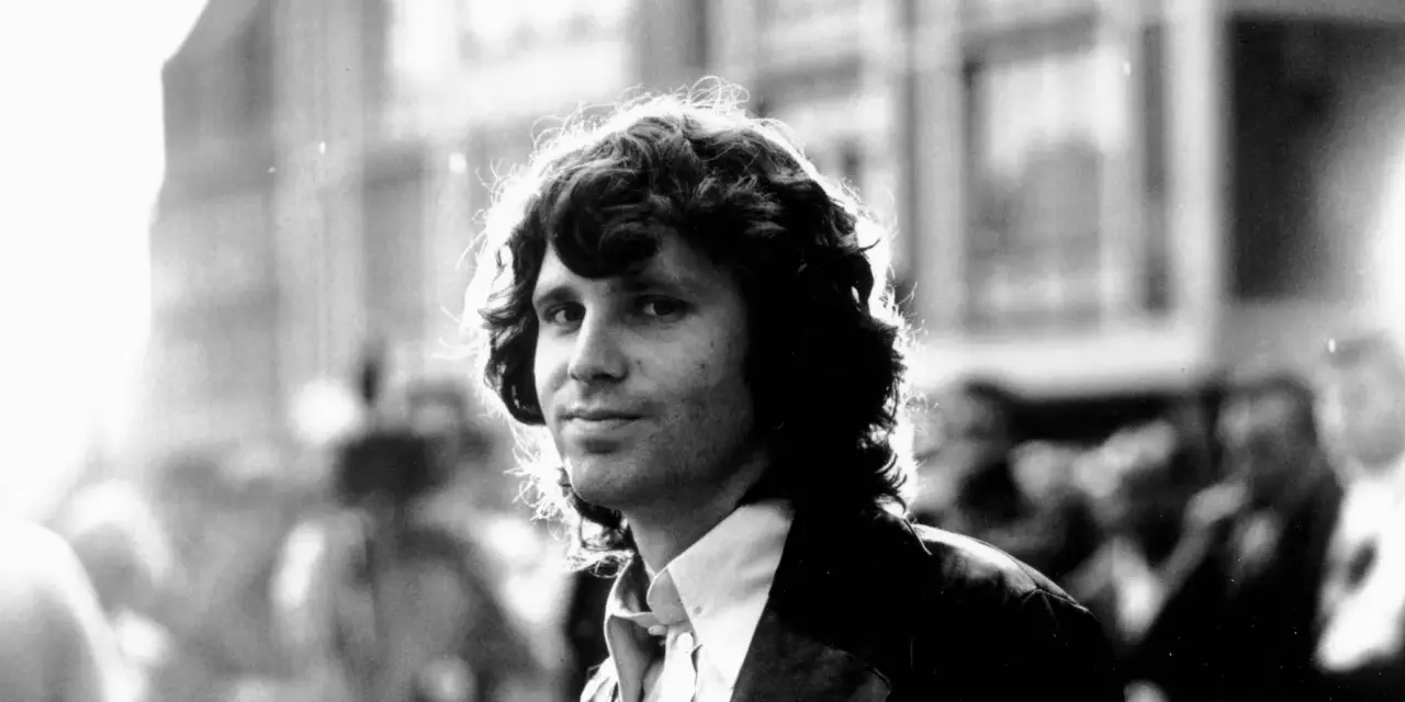 Jim Morrison, mit seinem charakteristischen lockigen Haar und einem nachdenklichen Blick, steht im Gegenlicht.