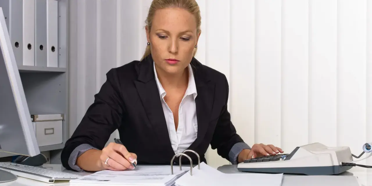 Kobieta w biurze pracuje nad dokumentami, używając kalkulatora i długopisu. Na biurku leżą stosy papierów i segregatorów, a w tle widać regał z dokumentacją. Może przygotowuje weksle.