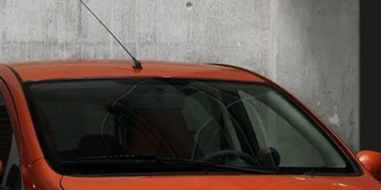 Pomarańczowy samochód z przyciemnianymi szybami z przodu, z antenką na dachu, stoi przy betonowej ścianie.