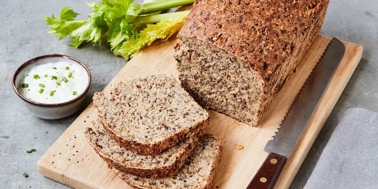 Ein rustikales Chia Brot, frisch aufgeschnitten, mit Sellerie und Dip auf einem Holzbrett.