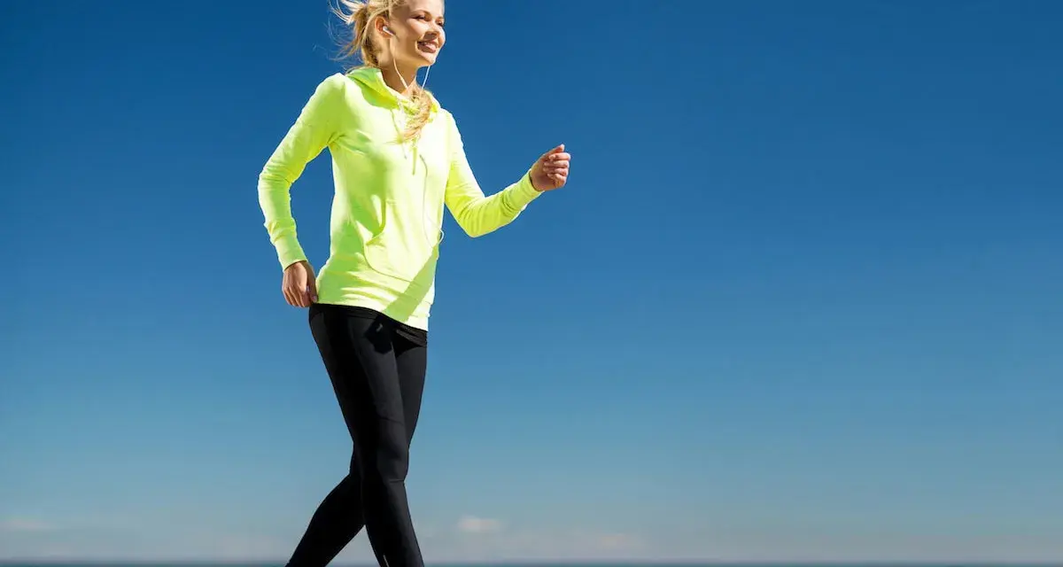 Uśmiechnięta blondynka w neonowej bluzie i czarnych legginsach uprawia power walking nad morzem.