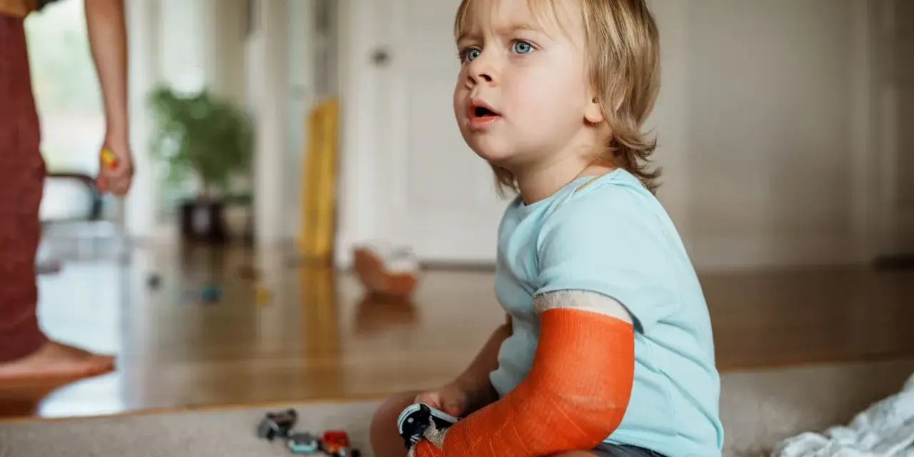 Kleiner Junge mit einem orangefarbenen Gips am Arm, der nachdenklich auf den Boden schaut. Er hat einen **kind arm gebrochen**.