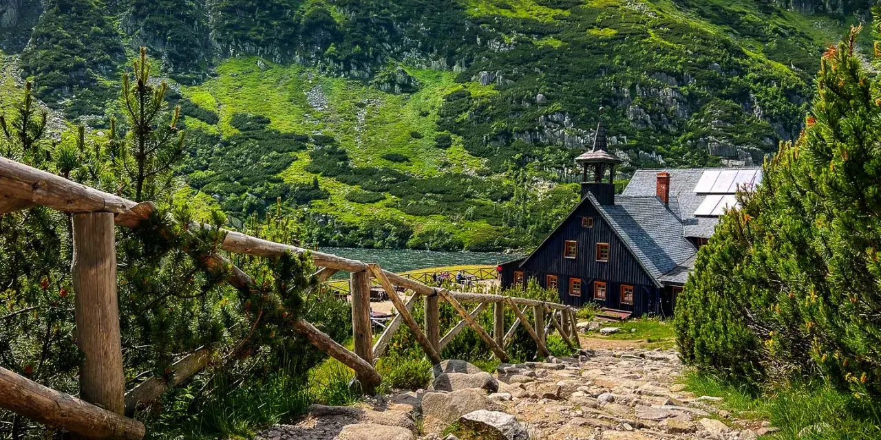 Kamienna ścieżka prowadzi do schroniska Samotnia Śnieżka nad jeziorem, otoczonego zielonymi zboczami gór.